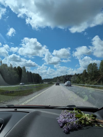 The image depicts a highway scene in Heinola, Finland, captured from the interior of a vehicle looking forward through the windshield. The foreground shows the top of a dark gray dashboard, with a small bunch of purple flowers, likely lilac, resting on its right side. A black windshield wiper base is visible at the base of the windshield.

The multi-lane asphalt highway extends into the distance under a bright blue sky filled with numerous white cumulus clouds. Sunlight causes a visible glare or lens flare in the upper left quadrant of the sky. Several vehicles are present on the highway. In the same direction of travel, a dark sedan is visible in the left lane, and a white panel van with an open rear door or trunk is in the right lane, followed by another dark vehicle further ahead. A light-colored car is seen traveling on the opposite side of the highway in the distance.

Both sides of the highway are lined with dense green deciduous and coniferous forests. The median or shoulder on the left side features green grass interspersed with patches of purple wildflowers, consistent with lupines. A bridge or overpass structure is visible spanning the highway in the middle distance. The road appears dry, and the overall scene indicates a clear, sunny daytime environment. Reflections of the car's interior and light sources are present on the windshield.