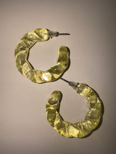 The image shows a pair of yellow, translucent, hoop earrings with a twisted design. The earrings are made of a material that appears to be acrylic or resin, with visible internal crackling or inclusions. Each earring has a silver post and a clear plastic backing. One earring is positioned to the left of the frame, with its post pointing upwards and slightly to the left. The other earring is to the right and slightly above the first, with its post pointing downwards and slightly to the right. The earrings are lying on a textured, light-colored surface that resembles leather or a similar material. The lighting in the image creates subtle shadows and highlights on the earrings, emphasizing their translucent quality and twisted form. There is no text visible in the image. The setting is a flat surface, likely a tabletop or display area. The image is a close-up, top-down view of the jewelry.