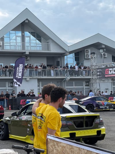The image, taken vertically, depicts a scene at the Rustavi International Motorpark in Rustavi, Georgia. The backdrop features a two-story building with a distinctive peaked roofline, numerous windows, and several air conditioning units mounted on the exterior.
Crowds of people are observing from the balconies and walkways of the building. Banners with sponsor logos like "GDC GEORGIAN DRIFT CHAMPIONSHIP" are displayed. There is also a flag with the words "ENERGIA BOLDIA".
Two race cars are prominently featured in the foreground. One is painted in dark green and yellow, with various sponsor decals, including logos such as "Valvoline" and "GT Garage" visible on the second, purple and yellow car.
A man wearing a yellow t-shirt is standing near the front car.