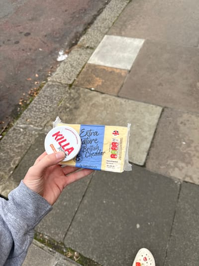 A close-up, high-angle outdoor shot depicts a person's left hand holding two packaged items over a flagstone pavement. The hand, with fair skin and short nails, emerges from a grey long-sleeved garment, likely a sweatshirt sleeve.
The primary item is a rectangular, plastic-wrapped block of "Extra Mature British Cheddar" cheese, branded "by Sainsbury's." Its label is blue and yellow, displaying a Union Jack flag, nutritional information, and a "Best Before: 06 JAN 26" date. Resting on top of the cheese block is a smaller, white, circular tub with a red lid. This tub is labeled "KILLA EXCLUSIVE NICPODS ORANGE ICE" and prominently displays the number "5," indicating nicotine strength.
The background consists of a worn, irregularly-laid flagstone pavement, featuring various shades of grey and brown, with some lighter square sections and visible cracks. Along the left edge of the pavement, a curbstone separates it from a darker, rougher ground surface, possibly a road or dirt verge, where small patches of green vegetation and detritus are present. The tip of a white shoe, possibly a Croc with red detailing, is partially visible in the bottom right corner of the frame.
The specific location within the city of Newcastle upon Tyne is not identifiable from the visual content of the image.