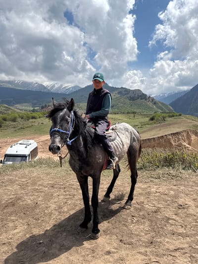 A person is depicted riding a dark grey or black horse in a scenic mountainous landscape in Jeti-Ögüz, Kyrgyzstan. The image is brightly lit, with strong shadows indicating midday or early afternoon.
The central subject is an individual wearing a blue cap, a dark vest, and a light-colored shirt, seated on the horse. The horse is fitted with a saddle and a blue bridle and reins, with its head turned slightly towards the viewer. The rider appears to be resting or paused on a dirt path.
In the background, majestic snow-capped mountains rise under a partly cloudy sky. The lower slopes of the mountains are covered with green vegetation. The midground consists of rolling hills with brown and sparse green growth. A patch of yellow flowers is visible on a bank to the right. The sky above is a clear blue with scattered white cumulus clouds.
Partially visible behind the horse, to the lower right, is a white vehicle, possibly an SUV or camper van, with the word "INTO" visible on its side. The scene conveys a sense of outdoor adventure amidst rugged natural beauty.