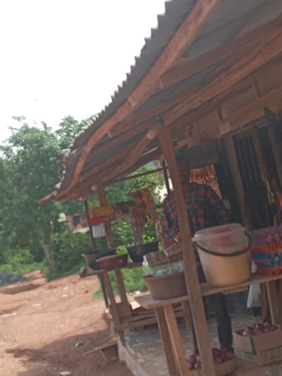 The image shows a roadside market stall in Achara Ndiagu Omegio, Nigeria. The stall is constructed of wood and corrugated metal. A person wearing a plaid shirt is visible behind the counter. Goods for sale, including onions, are displayed on shelves. Trees and vegetation are visible in the background. The ground is red earth.