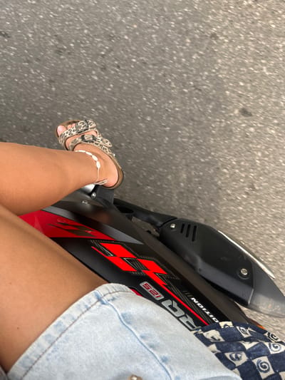 The image shows a close-up view of a person's legs and feet, resting on the seat of a motorcycle. The person is wearing leopard-print sandals and a denim skirt, with a shell anklet. The motorcycle is black and red, with "RR 125 EDITION" written on its side. The background is a blurry asphalt road, suggesting the motorcycle is in motion. The location is identified as Walahanduwa, Sri Lanka. The lighting indicates it is daytime.
