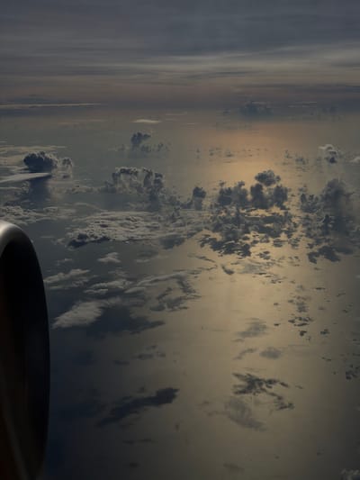 The image shows a view from an airplane window, likely taken at dawn or dusk. A portion of the airplane's engine is visible in the lower right corner. The sky is layered with clouds and a hazy atmosphere. The environment suggests the location is in the vicinity of Taoyuan City, Taiwan due to the typical weather patterns. There are no discernible people or specific actions.