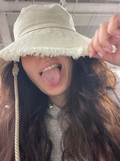 A person with long brown hair is seen from an upside-down perspective, wearing a light-colored hat with a frayed edge. Their tongue is sticking out. The hat has a patterned string attached. The background shows white structural elements and possibly the roof of a building. There is no specific information to confirm that this image was taken in Саларьево, Russia.
