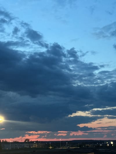 A low-angle, vertical, outdoor, eye-level photograph captures a wide expanse of the sky over a residential area at dusk. The sky transitions from a deep blue at the top to a hazy pink and orange near the horizon. Dark, heavy clouds dominate the upper and mid-sections of the frame, obscuring much of the lighter sky. Below the clouds, a faint, glowing light emanates from the horizon. In the lower portion of the image, a silhouetted landscape features the outlines of buildings, trees, and streetlights. A bright yellow light source, possibly a streetlight, is visible on the left side of the frame, casting a beam across the dark clouds. The scene appears to be located in Stony Plain, Canada, given the presence of typical Canadian residential architecture and vegetation under a vast sky. There are no people or animals visible in the frame.