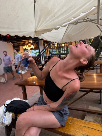 A Caucasian woman with blonde hair tied in a bun is sitting on a wooden bench under a large white umbrella. She is wearing a black crop top and grey shorts. Her head is tilted back, and she is drinking water from a clear plastic bottle held in her right hand. Water is dripping from her chin. She has tattoos on her left arm. To her left on the bench are a black backpack and a white and blue jacket. In the background, two men are walking. The scene appears to be outdoors at dusk, possibly at a restaurant or cafe, with wooden tables and chairs visible. Lights are visible in the background, suggesting artificial illumination. The ground is tiled with a reddish-brown color. The overall environment suggests a casual outdoor setting in a tropical or subtropical climate, consistent with a location like Cổ Lũng Commune, Vietnam.