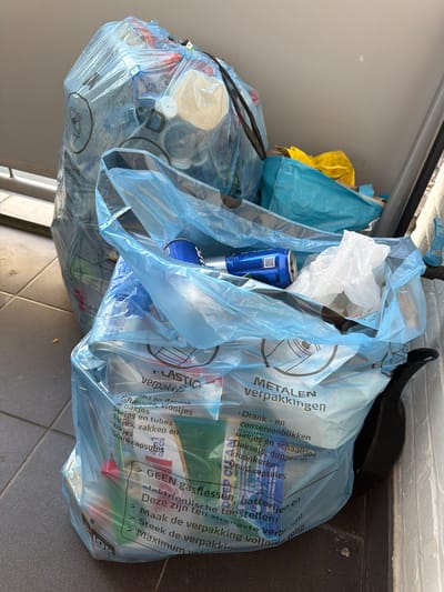 The image shows several translucent blue plastic recycling bags filled with various recyclable materials. One large bag in the background is packed tightly, with its opening pulled shut. A second, partially opened blue bag in the foreground contains several aluminum cans and some crumpled white plastic. Another blue bag, partially obscured, is behind the foreground bag. To the right, a yellow plastic bag is visible, peeking out from behind the larger blue bag. The bags are situated on a gray tiled floor, possibly an outdoor patio or balcony, adjacent to a white painted wall and a dark gray metal railing. The context of Turnhout, Belgium is not explicitly evident from the image itself, as there are no discernible landmarks or signs that specifically identify this location. The recycling bags appear to be awaiting collection or have been recently placed there.