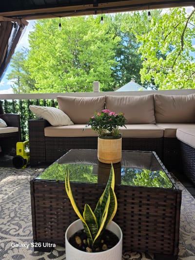 The image depicts an outdoor patio area, likely in West Hartford, Connecticut, United States of America, furnished with wicker furniture. A sectional sofa with beige cushions and a patterned throw pillow is visible, along with a wicker coffee table with a glass top. On the coffee table, a potted plant with small pink flowers sits next to a snake plant in a white pot, which is in the foreground. A neon yellow leaf blower is positioned on the floor to the left of the sofa. The background features lush green trees and a bright blue sky, suggesting a daytime setting. String lights are suspended above the patio, and a decorative green vine fence is seen along the railing. The ground is covered by a patterned outdoor rug. The scene conveys a relaxed, domestic outdoor living space.