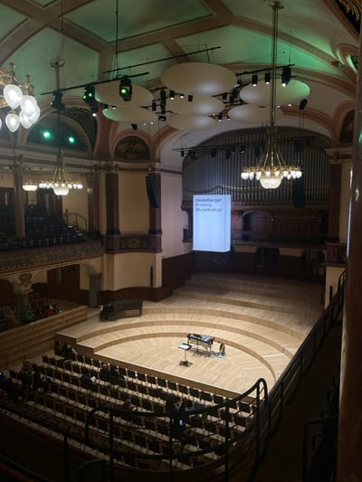 This image depicts the grand concert hall of the Stadthalle Heidelberg, Germany, viewed from an elevated position, likely an upper balcony. The scene showcases an ornate interior with a high, arched ceiling painted in pale green and cream, featuring decorative molding. Several elaborate brass chandeliers with multiple glass globes illuminate the space, complemented by modern stage lighting fixtures, some emitting green light, and large, oval acoustic reflectors suspended from the ceiling.

A large pipe organ dominates the rear wall of the stage area, below which a rectangular, illuminated white banner hangs, displaying "Heidelberger Frühling Musikfestival" in black text. The stage itself is semi-circular and tiered, constructed from light-colored wood. On the front section of the stage, a vibraphone or marimba, a music stand, and other percussion instruments are set up. Further back on an elevated tier of the stage, a black grand piano is positioned.

The audience seating consists of numerous rows of dark-colored chairs arranged on the main floor and in multiple tiered balconies with decorative railings. Scattered individuals are seated throughout the audience sections, appearing to be patrons observing or waiting for an event. Large dark speakers are mounted on the walls flanking the stage. Reddish-brown architectural pillars with ornate capitals are also visible within the hall.