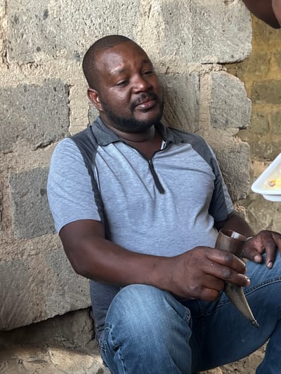 The image depicts a man sitting on a set of stone steps, likely located in a residential area of Githurai, Kenya, based on the provided coordinates. He is wearing a grey t-shirt and blue jeans, with his right leg bent and his left leg extended. He holds a small, dark object in his right hand and a larger, light-colored object in his left hand. The man's face is visible, and he appears to be looking down at the objects in his hands. The steps are made of rough, uneven stone, and there is some dirt and debris visible on the surface. The overall tone of the image is casual and informal, suggesting a relaxed and everyday setting.