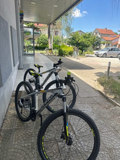 The image depicts a street scene in Ovoshtnik, Bulgaria. In the foreground, two mountain bikes with grey frames and yellow accents are parked on a tiled sidewalk. The bikes are positioned side-by-side, with the front bike partially obscuring the rear one. Behind the bikes, a building with a light-colored facade and a covered walkway is visible. Further in the background, a silver car is parked on a paved area, and residential buildings with red-tiled roofs are nestled amongst lush green trees. The sky is a clear blue with a few scattered white clouds. The scene suggests a sunny day in a residential area.