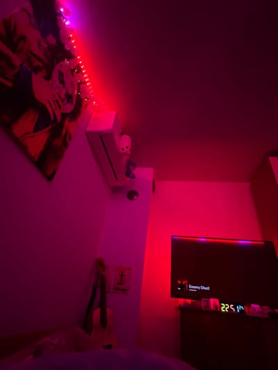 This interior scene, located in a room in Debelt, Bulgaria, is illuminated by predominantly red and purple LED lighting sources, casting magenta and deep red hues across the walls. On the upper left wall, a large framed artwork features a stylized depiction of a face with white makeup, resembling the Joker. A string of small, white flower-shaped lights is draped along the top edge of the artwork and extends to the right. Below these lights, a white rectangular console, similar to a Nintendo Wii, is mounted vertically on the wall. A small white figurine with a simple facial expression stands on top of the console, and a smaller blue figure is positioned directly below it. Further down the wall, a circular metallic fixture is visible. In the lower left foreground, a light-colored acoustic guitar leans against the wall. To its right, a small, square, light-colored object with a dark cross-like design is affixed to the wall. On the right side of the room, a flat-screen television is mounted, displaying a dark interface with the text "Enemy Diss!!" and "YungGoyardel". The television is bordered by red and purple backlighting. Below the television, a dark wooden dresser or stand supports several small, indistinct objects. A digital clock or display on the dresser shows green numbers "225" and blue numbers "124". The lower left foreground reveals the edge of a bed or cushioned surface. No individuals are present.