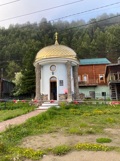 The image shows a small, light blue chapel with a golden dome topped by a cross, situated in Listvyanka, Russia. The chapel features stone pillars at its entrance and a set of white steps leading up to it. To the right of the chapel is a rustic wooden building with a green roof and balconies, and further to the right is another wooden structure. The foreground is a grassy area dotted with yellow wildflowers, and a stone path leads to the chapel. Behind the chapel, a dense forest of evergreen trees covers a misty hillside. Red cloths are tied to a fence in front of the chapel. A person in dark clothing is visible standing in the open doorway of the chapel. Overhead, power lines cross the frame.