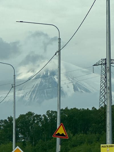 The image captures a landscape scene in the city of Svetly, Russia, dominated by a snow-capped mountain obscured by clouds in the background. In the foreground, a dense green forest occupies the middle ground. Several man-made structures are present: two tall, silver utility poles stand on either side of the frame, with power lines stretching between them and across the view of the mountain. A street lamp with a horizontal arm extends from the left pole. A triangular traffic sign with a yellow background and a black wavy line indicating a speed bump is positioned in the lower center of the image. To the far right, a portion of a metal lattice power pylon is visible. A yellow sign with the Russian word "МАГАЗИН" (store) is partially visible in the lower right corner. The overall lighting suggests an overcast day.