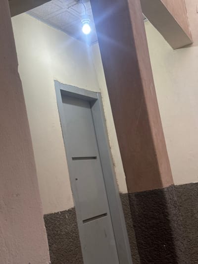 An upward shot reveals architectural details. A painted beam runs horizontally, casting a shadow. Below, a light shines against a wall where a rectangular vent with vertical slits is installed. The vent contrasts against the off-white wall. A portion of what appears to be a carpeted surface is visible along with a corner of a painted structure.