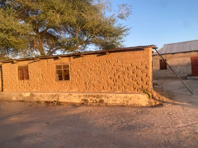 The image shows a mud brick building with two windows on the side, located in Dodoma, Tanzania, at the coordinates -6.177 latitude and 35.779 longitude. There is a tree in the foreground, a small concrete ledge in front of the building, and a dirt path leading to the left. The building has a corrugated metal roof and is partially obscured by a tree. The building appears to be an older structure. There is a second building in the background. The sky is clear blue, and there are no people visible in the image. The environment is a dusty and arid landscape, likely rural, with a dirt road in the foreground. The sun is setting and casting long shadows. The building appears to be in a state of disrepair, with some of the mud bricks crumbling. The building is likely a residential home or a small business. The scene suggests a simple life in a rural setting.