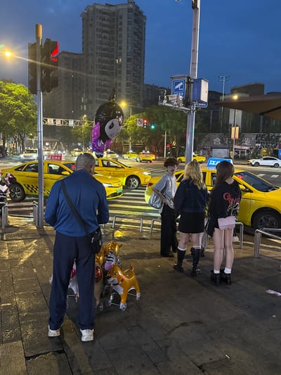 The image captures an outdoor street scene at dusk in Longta, China. In the foreground, a man in a dark blue shirt and pants stands facing away from the camera, with a black bag slung across his shoulder and another small bag at his waist. To his right, on a wheeled cart, are several inflated animal-shaped balloons, including a zebra and a dog.
Behind the man, a group of three young individuals are gathered near a yellow taxi. One person, wearing glasses and a grey shirt, stands to the left. To their right are two young women. The woman in the middle wears a dark denim jacket and a short black skirt, while the woman on the right is in a black hooded sweatshirt and a short pink skirt. They are all looking towards the street.
The street is populated with multiple yellow taxis, suggesting a busy urban environment. Traffic lights are visible, with one showing a red signal. Tall buildings line the background, indicating a city setting. A traffic camera and a surveillance sign are mounted on a pole to the left of the frame. The sky is a deep blue, transitioning into twilight. The ground appears wet, suggesting recent rain or street cleaning.