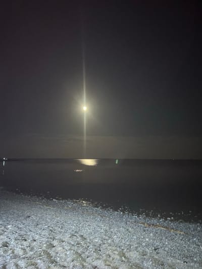 This is a nighttime photograph of a pebble beach and the sea under a moonlit sky. The moon is bright and positioned high in the sky, casting a reflection on the water. There are faint lights visible on the horizon across the water, possibly from distant buildings or boats. The foreground consists of a beach covered in small pebbles. The overall scene is dark and serene, with the moon being the dominant light source. The location is the shore of the Caspian Sea in the city of Karashygan, Kazakhstan.