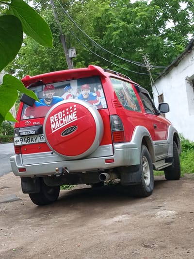 The image depicts the rear passenger side of a red Toyota Land Cruiser Prado parked on a dirt and asphalt surface. The vehicle is adorned with decals on its rear window, featuring images of hockey players, and a large "RED MACHINE" logo on its spare tire cover. A tiger decal is also visible on the passenger side window. In the background, a white building with a dark roof and a wooden structure are present. Green trees and foliage surround the scene, with power lines running overhead. The license plate of the vehicle reads "P948AY 12 RUS". The location is identified as Nakhodka, Russia.