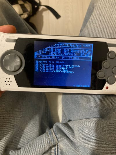 The image shows a close-up of a white handheld gaming device, likely a retro console, with a blue screen displaying a message about a missing drive. The device is held in a hand, partially obscured by blue denim jeans. The blue screen displays a series of text in a blue font, indicating a system error message. The text includes information about "Loader v1.46a," "Memory," "CPU," "Clock," and "TV NTSC." The screen also displays a message saying "No MD-Dos drive found." The device has a circular button on the bottom right side, a series of small holes around the bottom, and several small grey buttons near the top left corner. The text at the top right corner says "© 2017-2018." This device is being used in Kokhav Ya'ir Tzur Yigal, Israel, based on the provided coordinates.