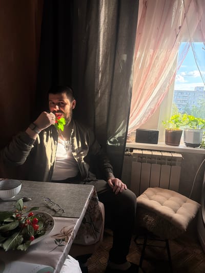 A medium shot captures a man with a beard and short hair sitting at a table, holding a cherry stem with leaves to his mouth. He is wearing a dark jacket over a white t-shirt, and a silver cross necklace. The table is covered with a floral-patterned tablecloth, and on it are a white bowl, a plate with cherries, and a pair of glasses. Sunlight streams through a window with sheer curtains, illuminating the scene and casting shadows. In the background, a radiator and potted plants are visible on a windowsill, with a cityscape visible through the window. The man is in a room that appears to be a residential apartment in Kolomna, Russia.