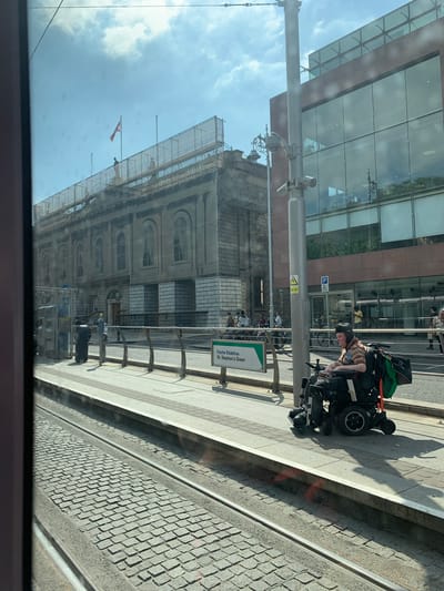 A person in a powered wheelchair is on the sidewalk near the light rail tracks in Dublin, Ireland. The location is the St. Stephen's Green station, evidenced by the station sign. The person is facing away from the camera. A large building is under construction and is situated behind the station sign, adjacent to a glass facade building. The photo appears to be taken from inside another vehicle.