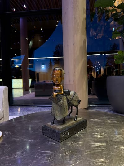A medium shot depicts a bronze statue of a seated figure with a fan, placed on a granite plinth on a grey-toned table. The statue is positioned near a large window reflecting an interior setting. A structural column is to the left of the window, and part of a plant is visible in the upper-left corner. The surrounding environment suggests an upscale interior, but there are no specific indicators to confirm the location in Озёрное, Russia.