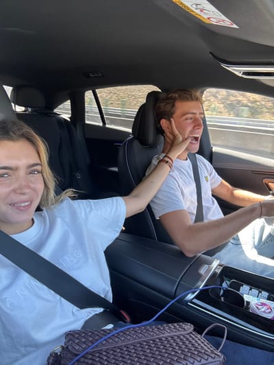 The image captures two young individuals inside a vehicle, appearing to be in motion on a highway or rural road. The background visible through the car windows shows a dry, hilly landscape with sparse vegetation and a road barrier, consistent with a region such as Pombal, Portugal.
In the passenger seat on the left, a female with light brown hair and a white t-shirt, bearing embossed text (partially visible as "ANING"), smiles directly at the camera. She is wearing a seatbelt, and a dark, woven shoulder bag rests on her lap. Her right arm is extended, with her open hand pressing against the left cheek of the male driver.
The male driver, positioned on the right, has light brown hair and is wearing a light-colored t-shirt with a small yellow logo on the left chest, along with blue jeans. He is secured by a seatbelt, with his left hand on the steering wheel. His mouth is open wide in an exaggerated expression, seemingly reacting to the female's touch. A brown leather bracelet is on his right wrist, and a red string bracelet is on his left.
The car's interior features dark upholstery, with the driver's seat having white piping. A modern center console with various controls and a visible blue charging cable separates the two individuals. An "AIRBAG" warning sticker is affixed to the passenger-side sun visor. The overall lighting suggests a sunny day.