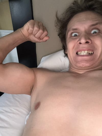 A close-up, chest-up selfie shows a shirtless person in bed, flexing their bicep and making a comical facial expression with wide eyes and gritted teeth. The person has messy brown hair and appears to be indoors, with a white pillow and a dark headboard visible in the background. The lighting suggests it is daytime. There is no indication of the location in San Francisco, United States of America.