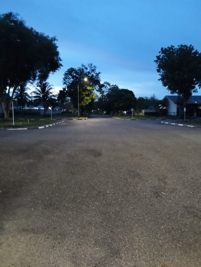 The image depicts an empty asphalt road in a suburban or campus-like setting at dusk. The sky is a deep blue, transitioning to a lighter hue near the horizon, suggesting the end of the day. A street light is illuminated on the right side of the road, casting a warm glow on the surrounding trees.
On the left side of the road, several trees, including palm trees, are visible. Further back, buildings with white walls and dark roofs are partially obscured by the trees. The road is lined with white and black markings, indicating a parking area or intersection.
The scene appears quiet and serene, with no people or vehicles present. The overall impression is one of peacefulness and solitude, typical of the early evening in a residential or institutional area. The location cues, such as the types of trees and buildings, and the consistent lighting conditions, suggest a tropical or subtropical environment. The image captures a moment of transition from day to night, with the natural light of the sky blending with the artificial light of the streetlamp.
