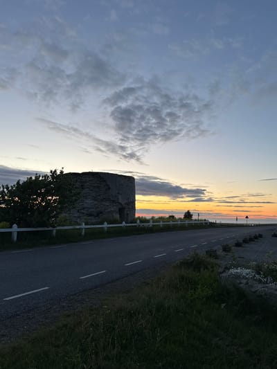 The image captures a panoramic view of a roadside in Paldiski, Estonia, at dusk. On the left side of the frame, a rustic, circular stone structure, possibly a historical ruin or a former military installation, stands prominently. Adjacent to it is a lush green tree, its silhouette illuminated by the fading light. A white fence runs along the roadside, separating the road from the area with the structure and vegetation.

The road itself is asphalt, with white dashed lines indicating lanes. The right side of the road features a gravelly shoulder with some potted plants and scattered stones. Beyond the road, a faint horizon suggests the presence of the sea, with a gradient of warm colors – oranges, yellows, and soft pinks – painting the sky, indicative of a sunset. Wispy clouds are scattered across the sky, with darker, more textured clouds present in the upper left portion of the image. The overall scene is tranquil and atmospheric, highlighting the unique landscape of Paldiski during the twilight hours. There are no people visible in the image.