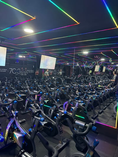 The image captures an indoor cycling studio located in Envigado, Colombia. The room features a dark ceiling and walls, significantly enhanced by an extensive grid of linear, multicolored LED strip lights that trace geometric patterns across the ceiling and along the mirrored back and side walls. A single oval-shaped recessed light is visible on the ceiling.

The studio is densely populated with numerous "Stages" brand stationary exercise bikes, arranged in rows. These bikes are primarily dark gray and black with blue accents, and some units feature integrated LED lighting that casts green and purple hues onto their frames. The floor is dark, likely covered with rubber matting.

The rear wall is composed of large mirrors, which reflect the bikes, lights, and people, creating a sense of depth. Motivational text and branding elements are visible on the wall behind the bikes, including "700 Style" and partial phrases like "you achieve" and "Dream without." Another mirror surface on the right displays stylized white text, including "Keep cool."

Two large rectangular screens are mounted on the mirrored wall. The left screen displays a bright, somewhat abstract image, while the right screen shows a digital interface with "WELCOME" text, indicating a login or interaction point.

Several individuals are present in the scene, primarily visible through reflections. On the left side of the reflections, two women are standing near bikes, one in a dark top and one in a lighter top. On the right side of the reflections, a man in a gray t-shirt is standing or adjusting a bike. In the physical foreground, on the far right of the image, a man with short hair, wearing a gray t-shirt and dark shorts, is seated on a stationary bike, facing away from the camera, appearing to be engaged in or preparing for a cycling session.