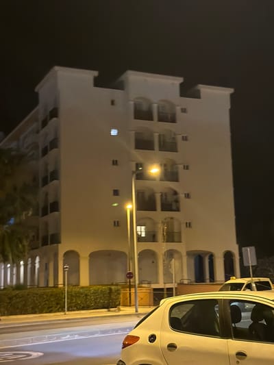 The image captures a nighttime street scene in Eivissa, Spain, dominated by a multi-story white building with arched balconies. The building has several illuminated windows and a prominent streetlamp casting a warm glow. In the foreground, the rear of a white car is visible, its taillight illuminated. A yellow taxi is parked behind the white car, partially obscured. A neatly trimmed hedge runs along the side of the building, separating it from the street. The sky is dark, indicating it is nighttime.