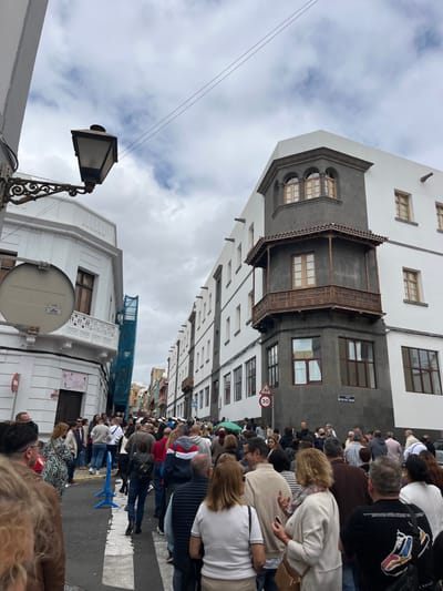 The image shows a street scene in the city of Las Palmas de Gran Canaria, Spain, located at coordinates 28.098 latitude and -15.416 longitude. The scene is crowded with people walking in both directions. The people are of various ages and ethnicities. Some are wearing casual clothing, while others are wearing more formal attire. Many are carrying bags and purses. There are buildings on both sides of the street. The buildings are mostly white with some gray accents. There is a streetlight on the left side of the street. The street is paved and has crosswalks painted on it. The sky is cloudy and the sun is not visible. The image was taken on a weekday afternoon.