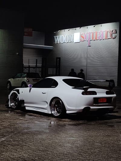 The image captures a white Toyota Supra parked in front of a building identified as "WOONsquare." The car features a prominent rear wing, aftermarket wheels, and a red license plate reading "39-HLD-2." A white Mini Cooper is parked behind it. Two silhouetted figures are standing near the building's exterior. The location appears to be at night, with artificial lighting illuminating the signage and the cars. The ground appears wet, reflecting the ambient light. The building has a corrugated metal exterior. Another sign with the text "SIC FIT" is visible near the entrance. The scene is likely in an urban environment based on the building design and infrastructure.