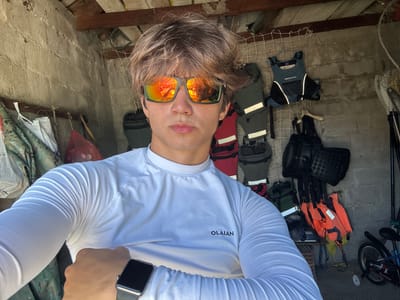 In Vranjina, Montenegro, a young male with light brown, tousled hair stands in an outdoor storage area. He is wearing a white, long-sleeved athletic shirt with the "OLAÏAN" logo visible on the front, and reflective, orange-tinted sunglasses. His left arm is bent and positioned across his chest, revealing a smartwatch on his wrist. Behind him, the scene is cluttered with various items including life vests of different colors (orange and dark blue), bags, a net, and other equipment, suggesting a space dedicated to outdoor or water activities. The wall appears to be constructed from concrete blocks, and the ceiling is made of wooden planks. A bicycle is visible in the background to the right.