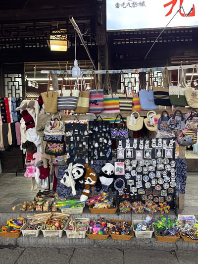 This image captures a street vendor stall in Pengshui Miao and Tujia Autonomous County, China. The stall is positioned against a backdrop of traditional dark wooden architecture featuring ornate latticework and a partially visible tiled roof. Overhead, a white sign with red Chinese characters is present, along with a modern white LED bulb and a traditional lantern hanging by wires.
The stall itself is densely packed with merchandise arranged on multiple levels. The upper level displays a variety of hanging bags, including straw totes, woven fabric bags in diverse colors and patterns (stripes, solids, multi-color crochet), and smaller decorative purses, some featuring embroidery. To the immediate left, various textile items like scarves, hats, and possibly gloves or small garments are neatly arranged on hangers.
The middle section of the stall presents numerous accessories. Several black display boards are covered with a multitude of earrings, ranging from simple studs to dangling designs. Below these, a collection of plush toys is visible, prominently featuring two black-and-white panda bears and what appears to be a red panda. Adjacent to these are additional small embroidered bags and various cultural souvenirs, including several rows of white ceramic cat-like figurines and multiple circular and rectangular pins or magnets depicting scenic or traditional motifs. A beaded necklace is also displayed.
The lowest level of the stall, resting directly on the stone-paved ground, consists of multiple white and brown wicker-like baskets filled with smaller items. These include miniature wooden toys such as trucks and construction vehicles, plastic toys, brightly colored hair accessories (e.g., scrunchies), small puzzles like Rubik's cubes, keychains, and assorted trinkets. No individuals are visible actively interacting with the stall or its merchandise, presenting the scene as a static display.
