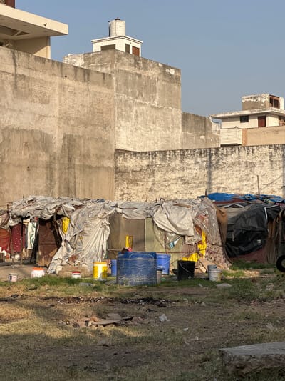 The image depicts a low-income area in Mathura, India, with dwellings constructed of tarps and assorted materials adjacent to unfinished concrete buildings. Piles of debris and discarded objects, including plastic buckets, paint cans, and textiles, are scattered along the edge of a dirt and grassy area. The buildings are multi-story, with exposed concrete facades. The sky is pale blue. The scene implies poverty and makeshift living conditions.