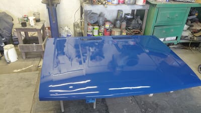 In an interior workshop setting, potentially located in Belovo, Russia, a freshly painted, high-gloss blue car hood is the central subject. The hood rests on a makeshift stand, with its surface reflecting the overhead lights and the surrounding clutter. To the left of the hood, a small, worn wooden stool holds several cups and containers, one of which appears to be a white ceramic mug. Next to the stool, a white metal pitcher with a handle is positioned on the concrete floor.

The background reveals a cluttered workshop environment. A shelf unit filled with various cans, bottles, and tools is visible behind the hood. To the right, a green metal cabinet with two drawers stands against the wall, also containing various items. Electrical cords and chains are scattered on the floor in front of the cabinet. The overall impression is one of a working garage or repair shop.