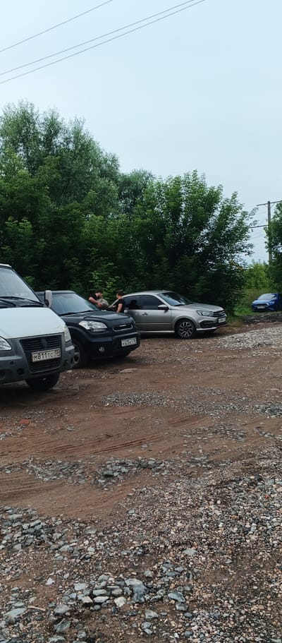 A light silver sedan, a black hatchback, and a white minivan are parked on a dirt and gravel lot. Two individuals are standing between the silver sedan and the black hatchback, appearing to interact with objects on or in the vehicles. A blue sedan is parked further down the lot. Dense green foliage borders the lot. Overhead, utility lines are visible against a light sky. The license plate of the white minivan is visible and reads "M811TC 18". The license plate of the black hatchback is visible and reads "K625KT 18". The license plate of the silver sedan is partially visible and appears to read "H496CE 18". The scene is located in the city of Pirogovo, Russia.