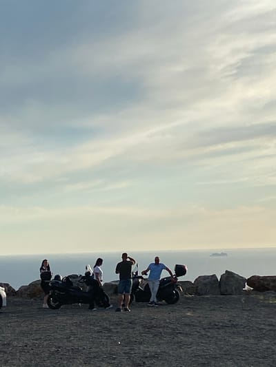 In the city of Kovačko Polje, Montenegro, on a rocky terrain overlooking a vast body of water, a group of four individuals are gathered around two motorcycles. The sky above is a soft blend of blues and whites, with faint clouds drifting across the horizon. The sea stretches out, meeting the sky in a hazy line, and a distant ship is visible on the water.
On the left, a woman with dark hair, wearing a dark top and shorts, stands near a black motorcycle. To her right, a second woman, with dark hair and wearing a white t-shirt and dark pants, leans against the same motorcycle. A man with dark hair, in a dark t-shirt and shorts, stands near the front of the motorcycle, with his hand near his head as if adjusting something or shielding his eyes.
Further to the right, a man in a light blue t-shirt and light-colored pants stands with one hand on the handlebars of a second black motorcycle. This motorcycle has a red storage box on the back. The man appears to be interacting with his companion or the motorcycle. The rocky landscape behind the people and motorcycles is uneven, with large, dark boulders scattered throughout. The ground in the foreground is a mix of dark gravel and small stones.