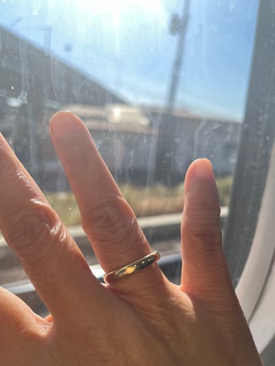 A close-up, over-the-shoulder view captures a hand wearing a simple gold band on its ring finger, positioned in front of a window. The hand, exhibiting clear wrinkles and texture, is illuminated by sunlight, casting shadows. The window is dirty, with streaks and smudges obscuring the view outside. Through the smudged glass, a blurred background suggests an outdoor environment with a blue sky and distant structures, possibly a street scene in Grengseng, Indonesia. The overall impression is one of everyday life, seen through a grimy lens.