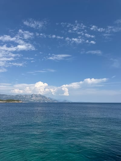 The image depicts a serene seascape with a view of the coast and mountains. The foreground is dominated by the calm, deep blue waters of the Adriatic Sea, transitioning to a lighter turquoise hue closer to the shore. Gentle ripples disturb the water's surface. In the midground, a rugged coastline is visible, dotted with sparse greenery and some scattered white structures, suggesting a settlement. Behind the coast, a range of majestic mountains rises, their peaks partially obscured by a scattering of white cumulus clouds against a bright blue sky. The clouds are dispersed, with some larger, more voluminous formations contrasting with thinner, wispy streaks. The scene is bathed in natural daylight, creating a peaceful and picturesque atmosphere. There are no people or discernible actions visible in the image. The location is identified as Dubraljevina, Montenegro, which is known for its coastal beauty and mountainous terrain.