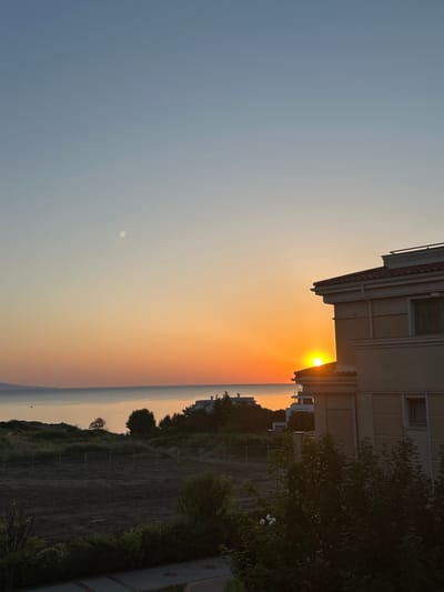 The image captures a sunrise over the Black Sea, viewed from Chernomorets, Bulgaria. The sun is low on the horizon, casting a warm orange glow across the sky and water. In the foreground, a modern, beige building with a red-tiled roof is partially visible on the right. Below the building and extending towards the sea, there is a patch of dark, tilled earth, bordered by a low wire fence. Further in the mid-ground, there are scattered trees and some smaller, multi-story buildings, indicative of a coastal town. The sea itself is calm, reflecting the colors of the sunrise, with a distant landmass visible on the left horizon. The sky transitions from a pale blue at the top to a vibrant orange near the horizon, with a faint, bright spot, possibly the moon or a bright cloud, in the upper left quadrant. The overall scene suggests an early morning coastal view.