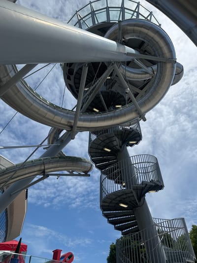This image captures a low-angle view of a large, metallic, spiral slide structure set against a bright blue sky with scattered clouds. The slide is made of gleaming silver material, with transparent sections revealing glimpses of the interior. Multiple circular staircases with metal railings spiral upwards, connecting to different levels of the slide. The stairs are illuminated by modern, rectangular lights.
In the background, to the left, a modern building with glass windows is visible. Below the slide structure, a red umbrella and a red sculpture that appears to spell "LOVE" are partially visible.
This structure is located at Aventura Mall in Aventura, Florida, United States of America, and is part of the mall's architectural design and recreational amenities. There are no people visible in the image.