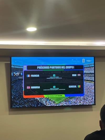 The image shows a television screen displaying upcoming matches from "Grupo I" of the FIFA World Cup 2026. The screen exhibits the VIX logo and lists two matches: "Francia vs Irak" and "Noruega vs Senegal". Both matches are scheduled for "Lunes 22 de Junio" (Monday, June 22nd). Details about the stadium and broadcast time are also visible, with one match listed as being at "Estadio Filadelfia | 2:45 PM for VIX" and the other at "Estadio Nueva York | 5:49 PM for VIX". The television is mounted on a wall, with a ceiling light visible above it. In the foreground, a person wearing a black shirt is seated and partially visible from behind, facing the television. The scene appears to be indoors. Based on a logo on the screen reading "VIX", it is highly probable that this is being viewed in Mexico, given that VIX is a major streaming service in that country. While the specific location within Cuautitlán Izcalli, Mexico cannot be definitively determined from the image alone, the presence of the VIX logo strongly suggests a viewing location within Mexico.
