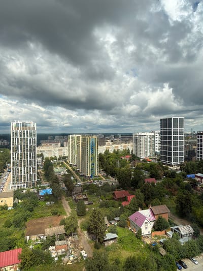This is an aerial, high-angle, medium shot of a developing urban landscape, likely within the city of Lyuk, Russia, under a cloudy sky. In the foreground, there are several older, smaller, and somewhat dilapidated residential buildings surrounded by dense greenery and trees. Some of these structures appear to be single-story homes with corrugated metal roofs, while others are larger, multi-story buildings with more modern, tiled roofs. There is evidence of construction and debris scattered around some of the older buildings, suggesting ongoing renovation or demolition.
Further into the middle ground, there is a park-like area with a sports court, likely a basketball court, visible. The park is bordered by trees and pathways, and several cars are parked nearby. Beyond the park, a cluster of modern, high-rise apartment buildings dominates the scene. These buildings are characterized by their geometric designs and the use of glass and concrete in their construction. One building on the left is tall and slender with a white exterior and numerous windows divided by green and white patterns. To its right, another building features a colorful facade with vertical bands of green, yellow, and blue. Further to the right, a dark gray building with a grid-like pattern of white window frames stands out. In the background, more apartment buildings and residential structures are visible, extending towards the horizon, which is lined with a dense forest. The overall impression is one of contrast between older, rural-style dwellings and newer, modern urban development, all situated within a lush, tree-filled environment. There are no people visible in the frame.