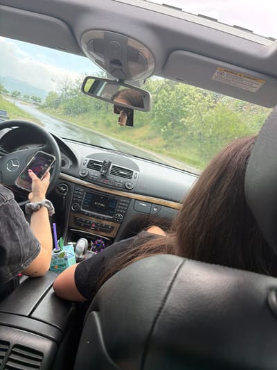 The image depicts the interior of a car, specifically the driver's side. A person is holding a mobile phone in their left hand, resting on the steering wheel. The car's dashboard is visible, displaying the radio and other controls. A drink container is in the center console area. A second person is sitting in the passenger seat. The scene appears to be on a road, visible through the windshield and side windows. The location information provided is Belo Pole, Bulgaria.