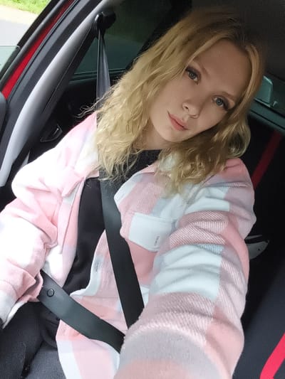 A blonde woman with blue eyes is sitting in the driver's seat of a red car. She is wearing a pink and white plaid shirt and a black t-shirt. A seatbelt is fastened across her chest. The interior of the car is visible, including the dashboard and a portion of the passenger seat. The woman is looking at the camera and smiling. Outside the car, greenery is visible. The image was taken in Plamya-3, Russia.